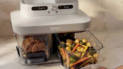 An air fryer with two glass baskets and a grey head unit on a marble kitchen counter. One basket contains meat, the other roasted vegetables