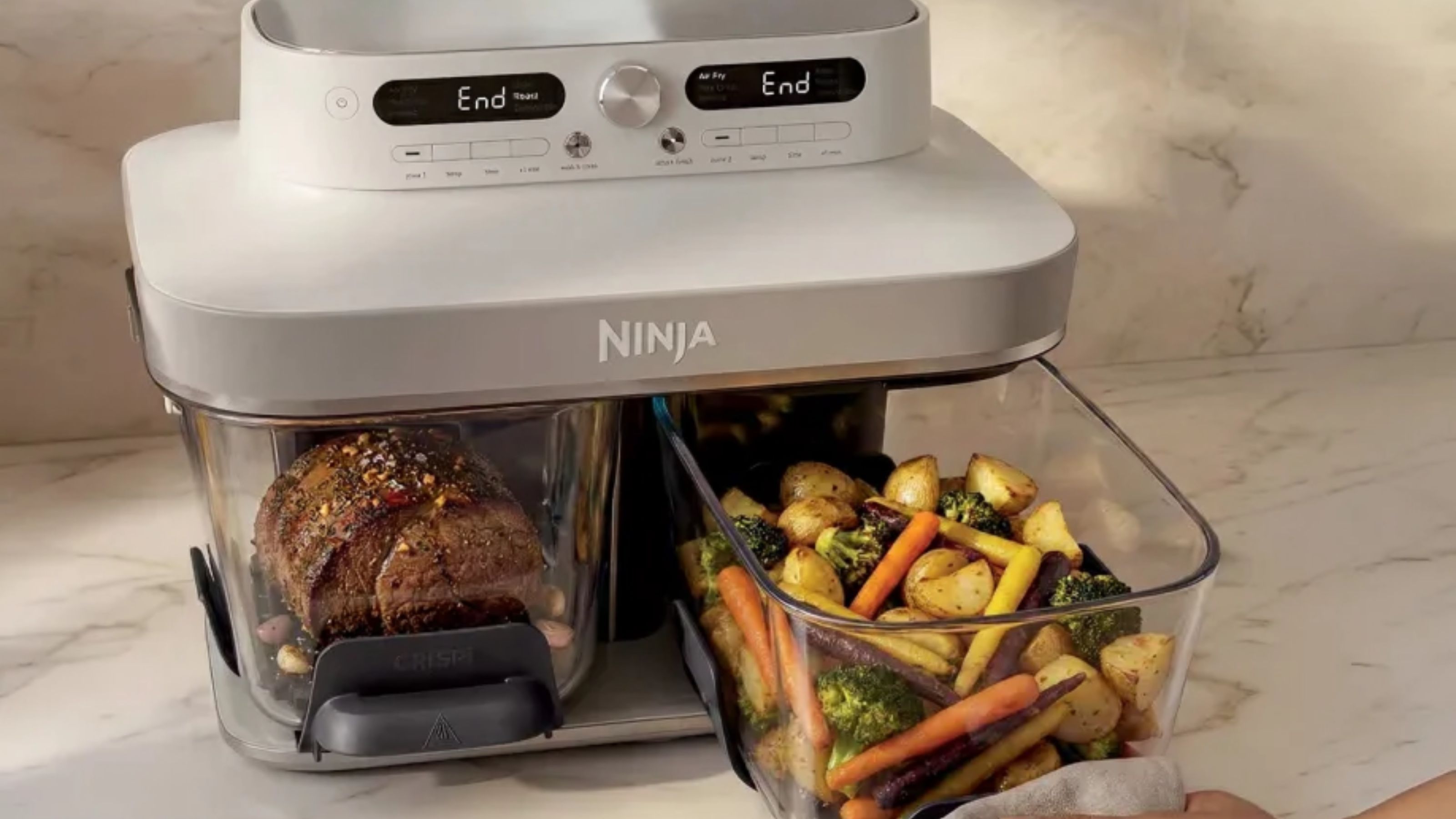 An air fryer with two glass baskets and a grey head unit on a marble kitchen counter. One basket contains meat, the other roasted vegetables