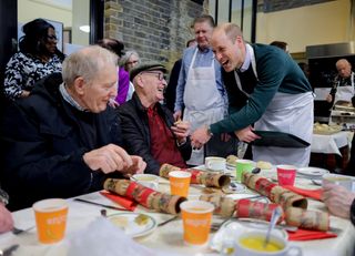 Prince William wearing an apron and laughing with two men sitting at a table full of plates and cups