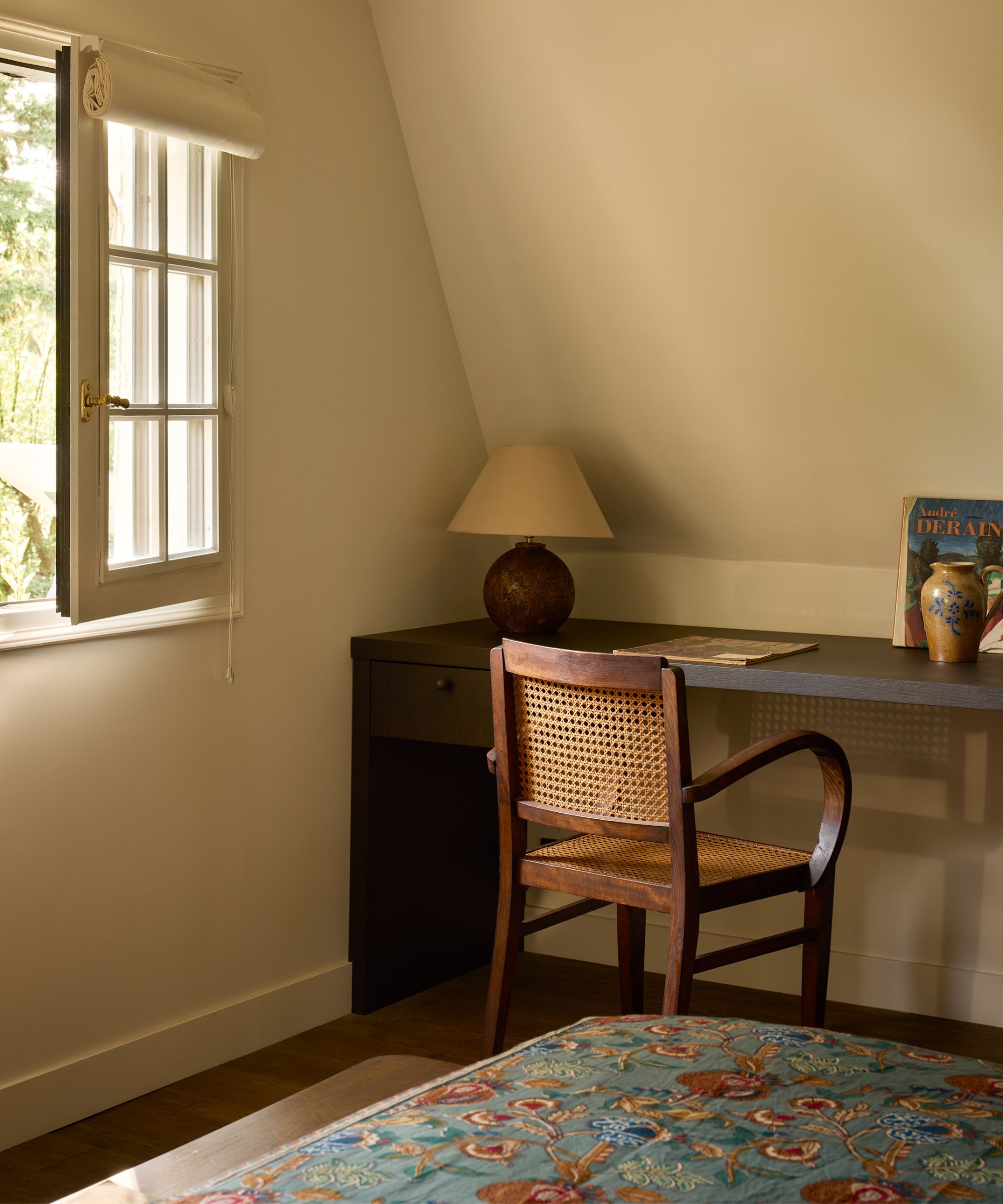 a light beige bedroom with a small window, dark wooden vintage desk, rattan chair, and a bed with a floral blue bedspread