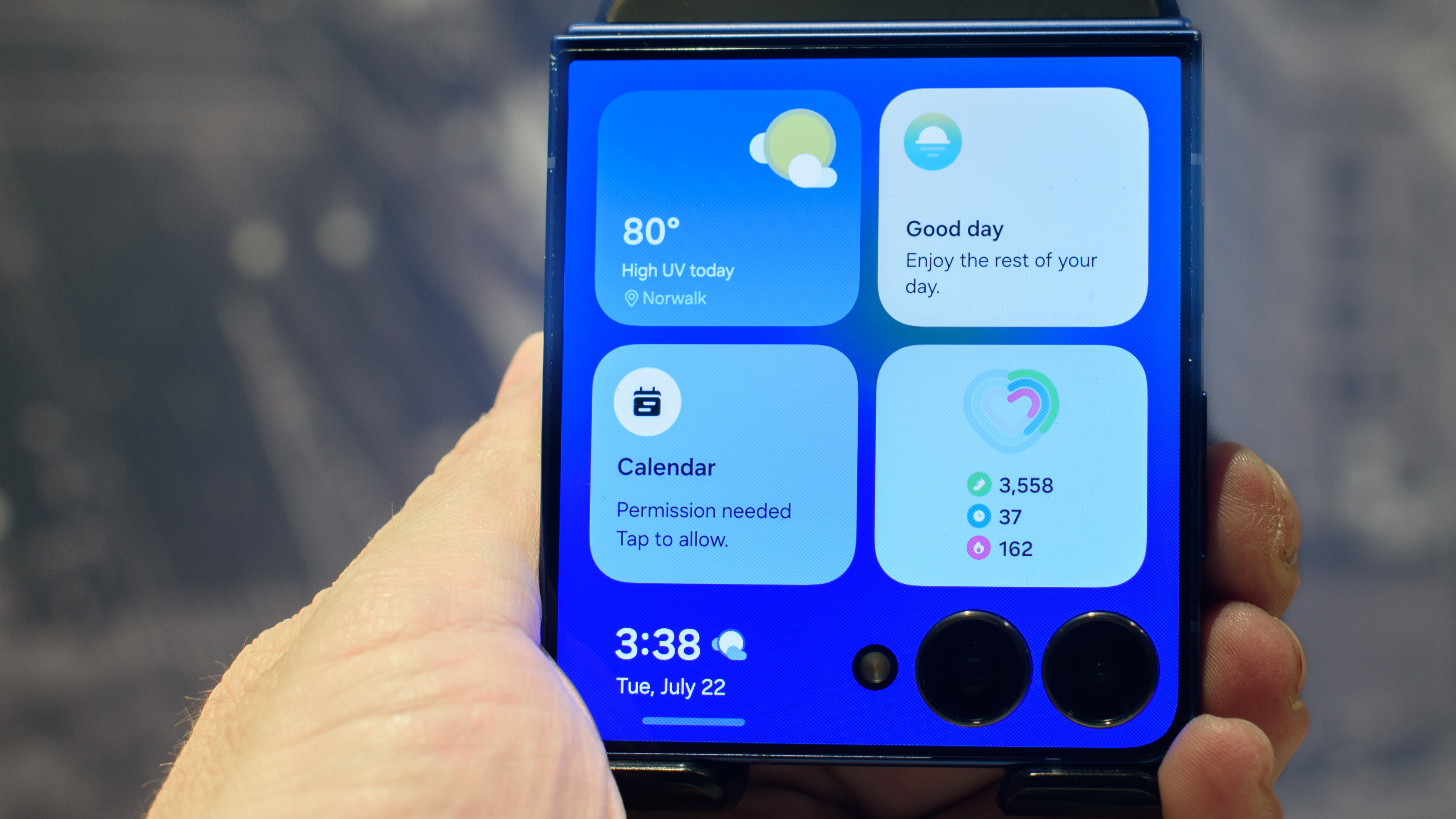 Samsung Galaxy Z Flip 7 in blue in hand showing four widgets on the cover display including weather, now brief, calendar, and health monitor
