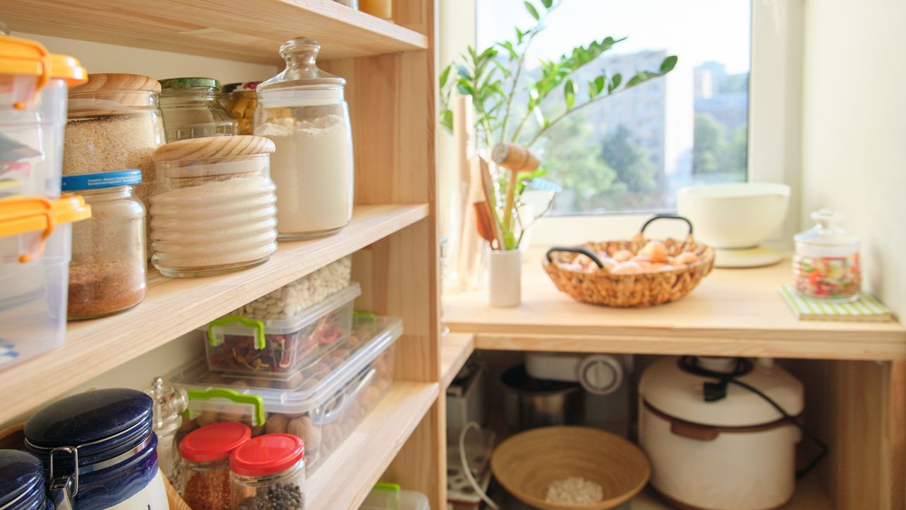 An image of a kitchen food pantry with wooden shelves and jars of dried goods stored on them and cooking appliances and other foods