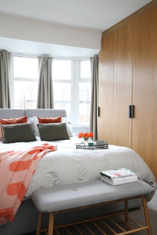 Bedroom with grey bed and bench, wooden fitted wardrobes, striped white and grey bedlinen and coral accessories