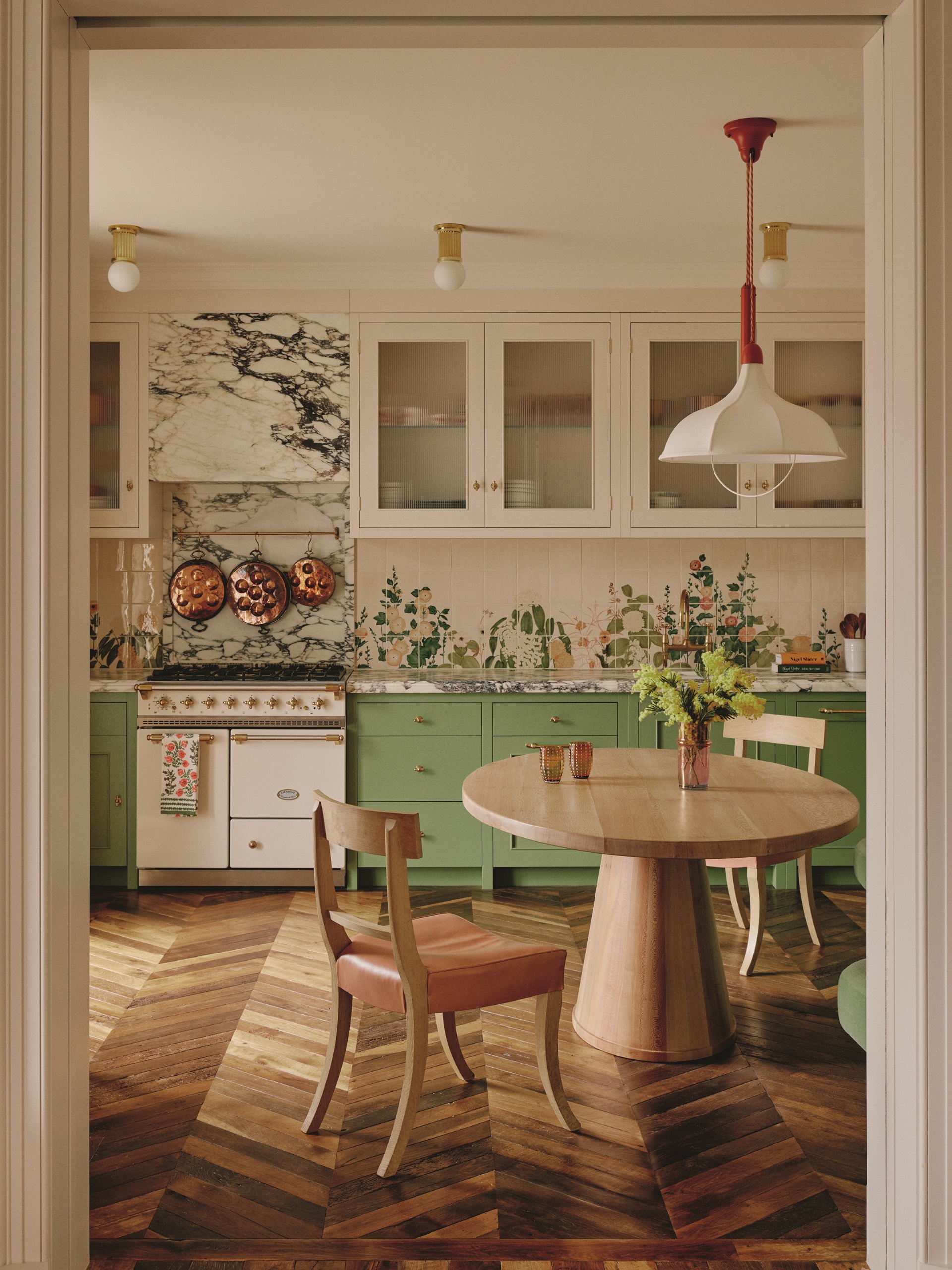 a green kitchen with parquetry floor, round table, timber dining chairs, marble over the cooker and a mural backsplash tile under white cabinets