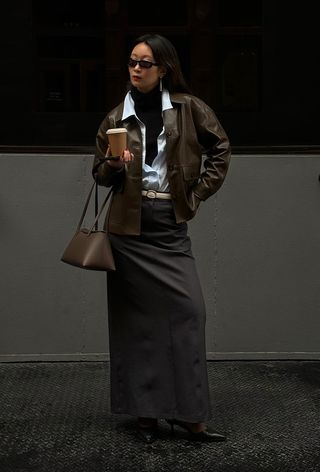 A cute work winter outfit is shown in a photo of a woman standing on the sidewalk in New York wearing a black turtleneck under a white button-down styled with a brown leather jacket, white belt, gray maxi skirt, black tights, black pumps, and a brown handbag