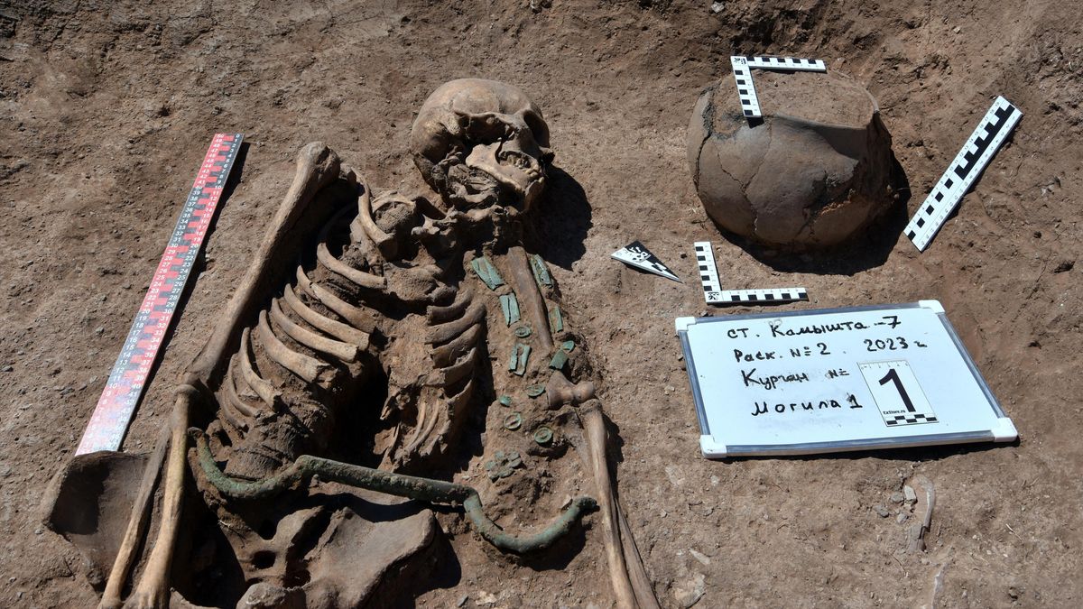 We see a skeleton in a dirt burial. There is a bronze bar with hooked ends across the skeleton&#039;s waist and a bowl to the upper right.