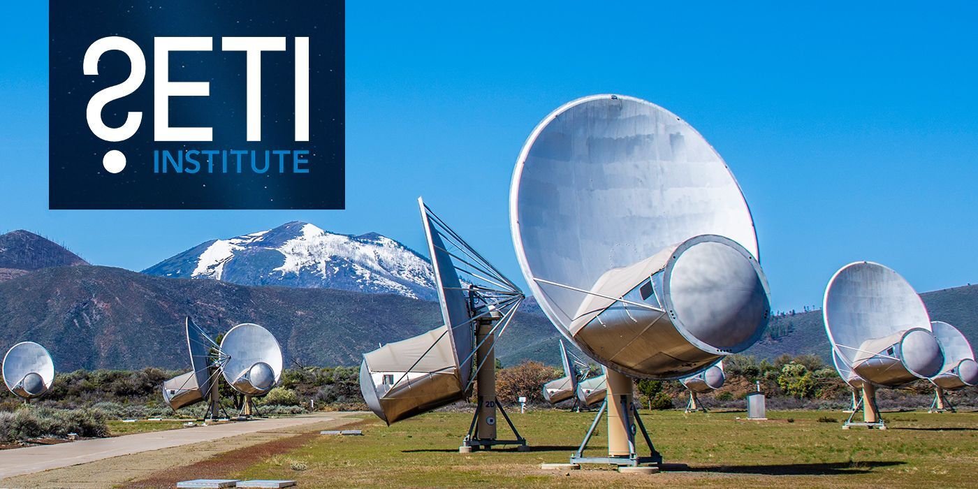 a large white satellite dish sits in a grassy field before a large snowing mountain in the background of a blue sky.