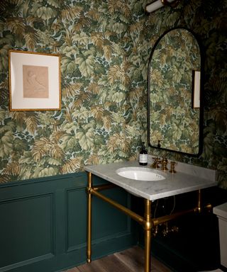 An example of powder room wall decor showing a dark green powder room with green botanical wallpaper and a traditional sink