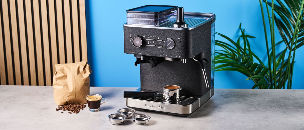 a black espresso machine by kitchenaid photographed against a blue background with silver chrome buttons and a pressure dial and steam wand with a tamped shot and a bag of whole coffee beans