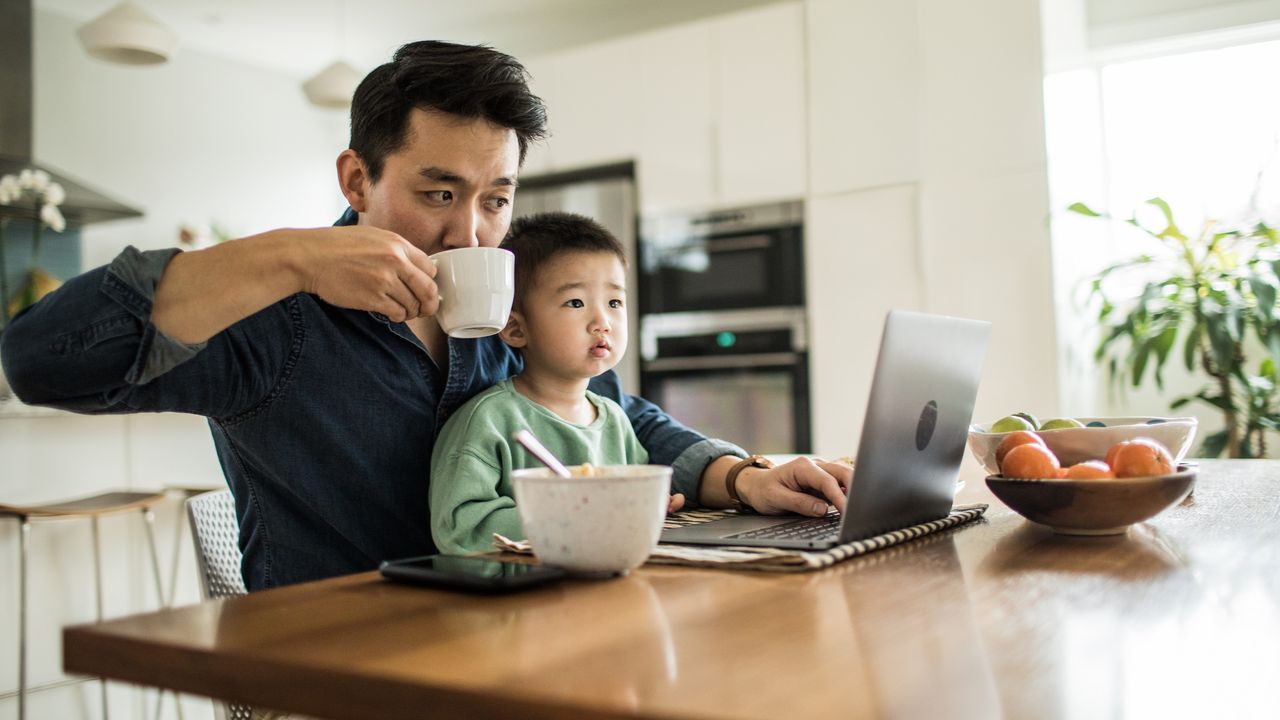 A father works on his laptop at the kitchen table while his young son sits on his lap