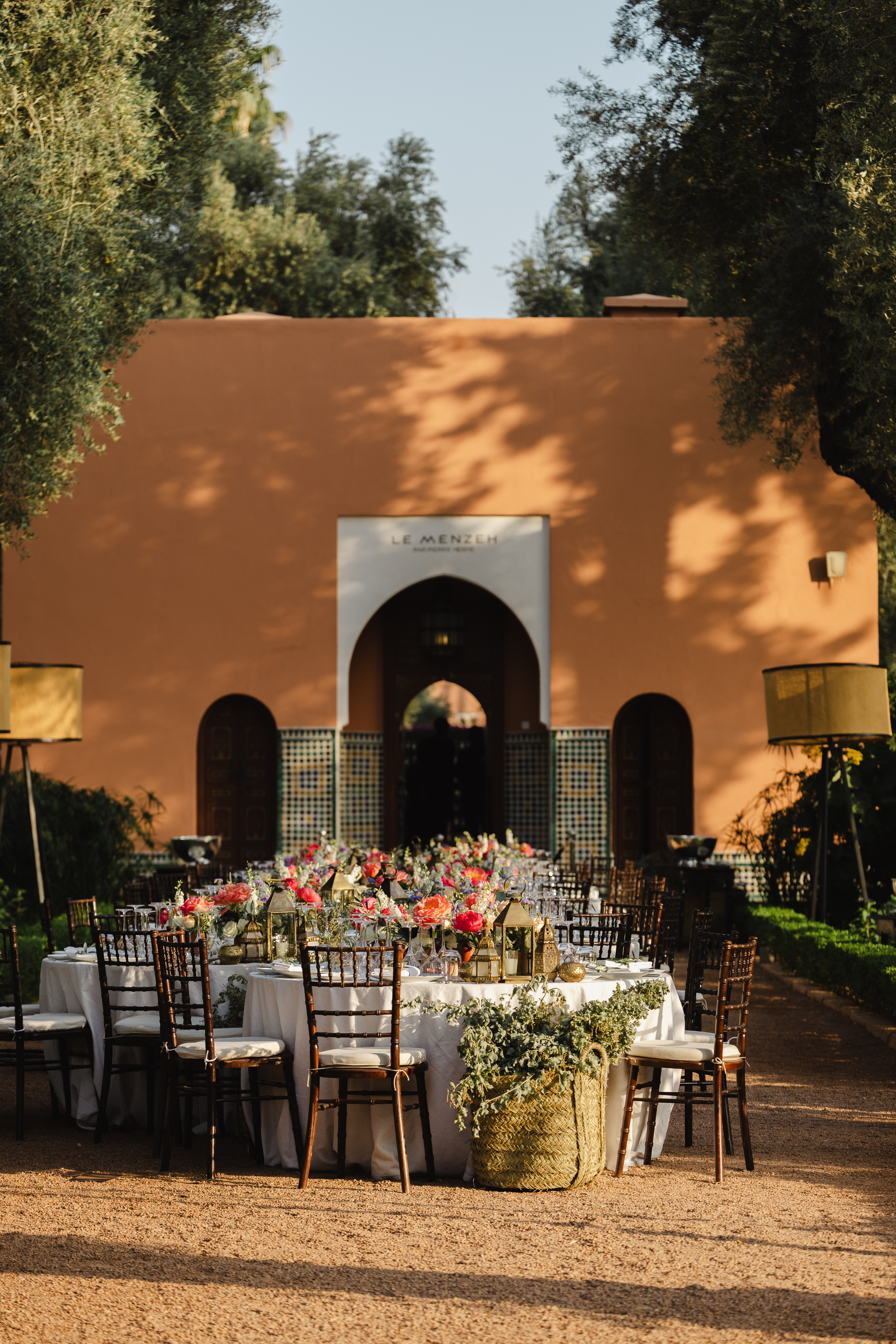 A sun-kissed table set in a garden punctuated by olive trees and ochre-tinted standing lamps, delineated by a terracotta arch with blue, green, and yellow tiles.