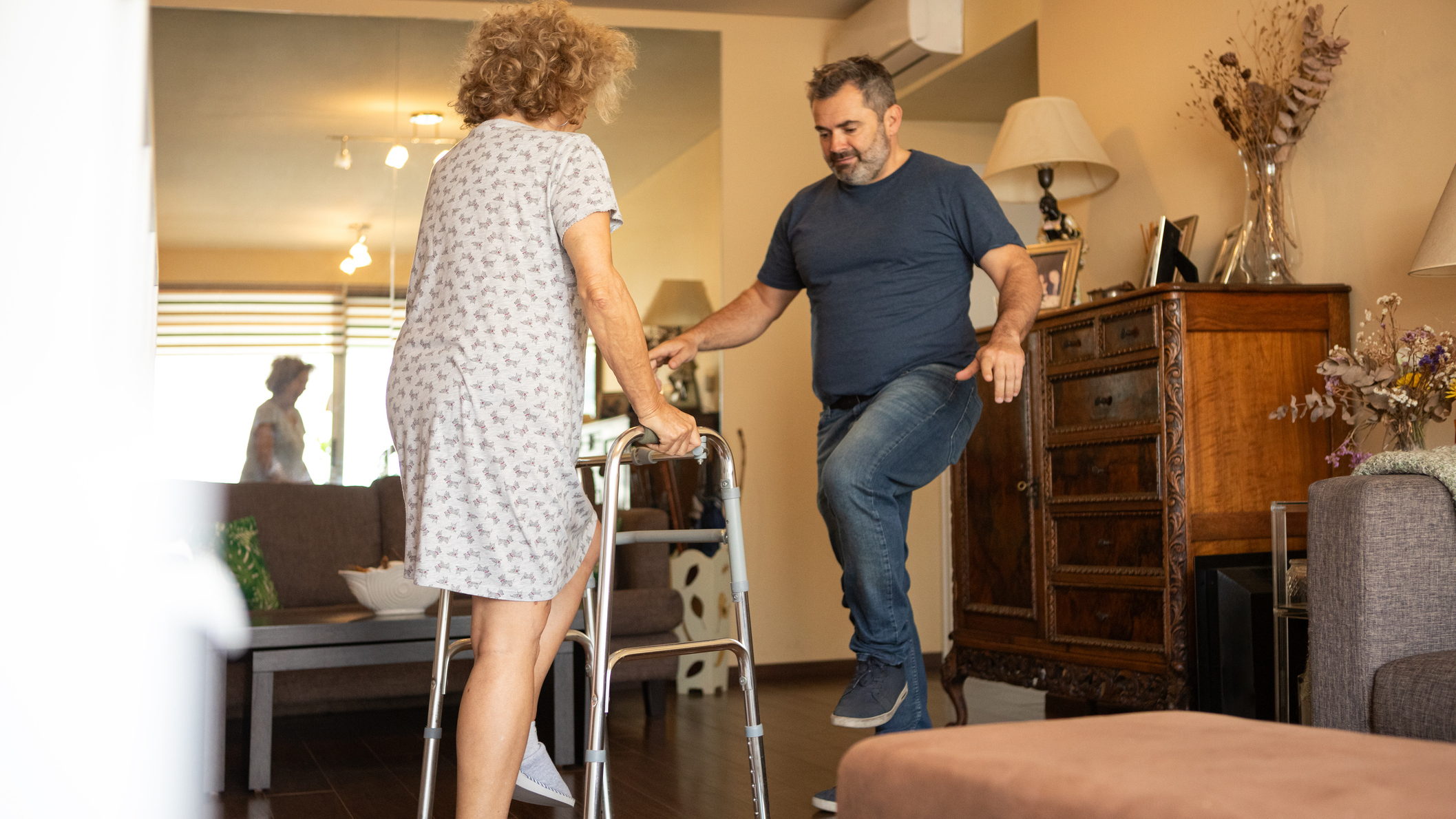 Woman in medical gown and walker faces man in jeans and T-shirt, they both lift one leg