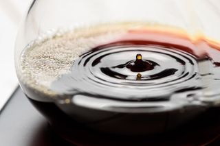 A jug of coffee with a drop of coffee hitting the liquid's surface, causing ripple of coffee. It is implied that this is coffee being prepared in the drip method