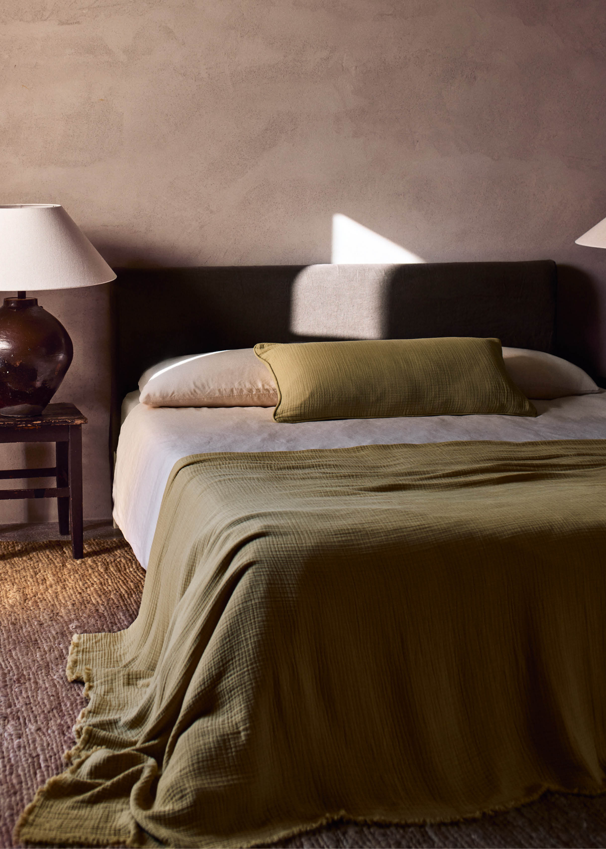 A bedroom with limewash walls, brown lamps with white shades, a textured area rug, and a gray bed with an olive accent pillow and throw
