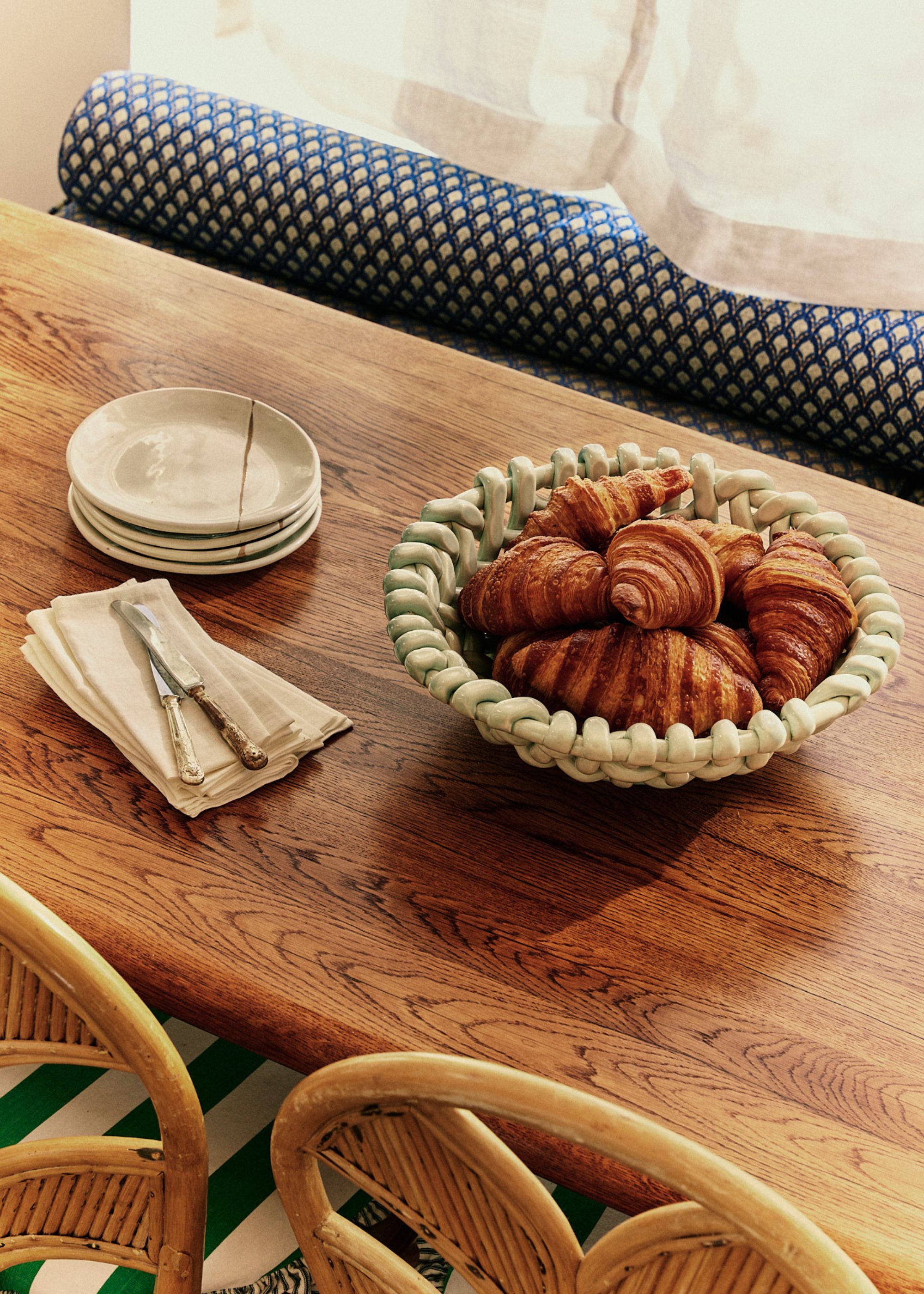 A white basketweave bowl with croissants inside of it. There are also a stack of side plates with butter knives and napkins on an oak table
