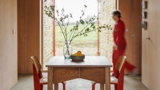 A wood-paneled living room with a central, square table, red-painted wooden chairs, a transparent glass vase, and a fruit bowl filled with oranges, looking out to greenery.
