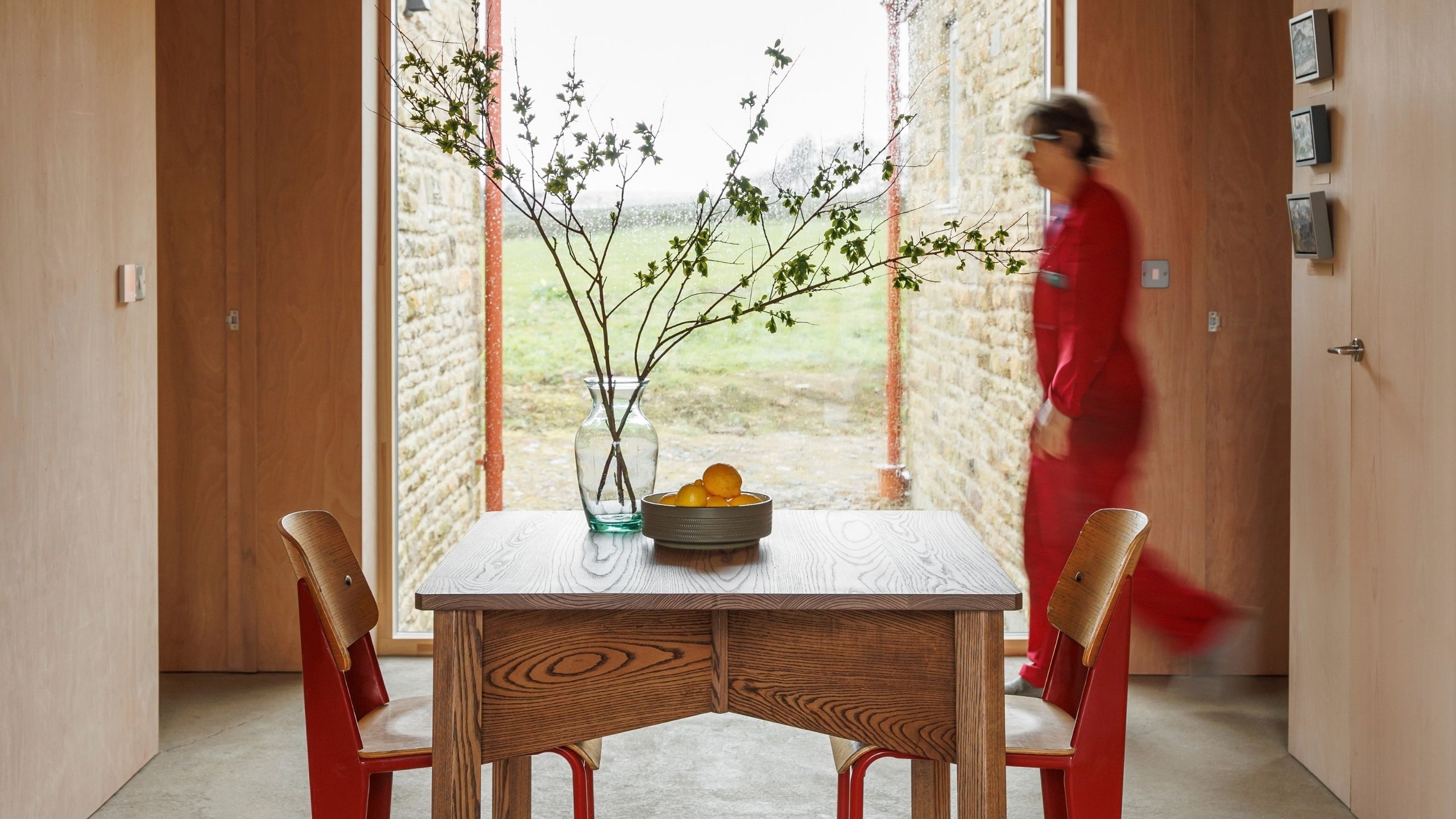 A wood-paneled living room with a central, square table, red-painted wooden chairs, a transparent glass vase, and a fruit bowl filled with oranges, looking out to greenery.