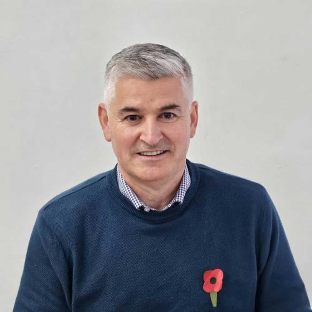 Smiling grey-haired man in a navy jumper with a poppy pin, headshot against a plain background.