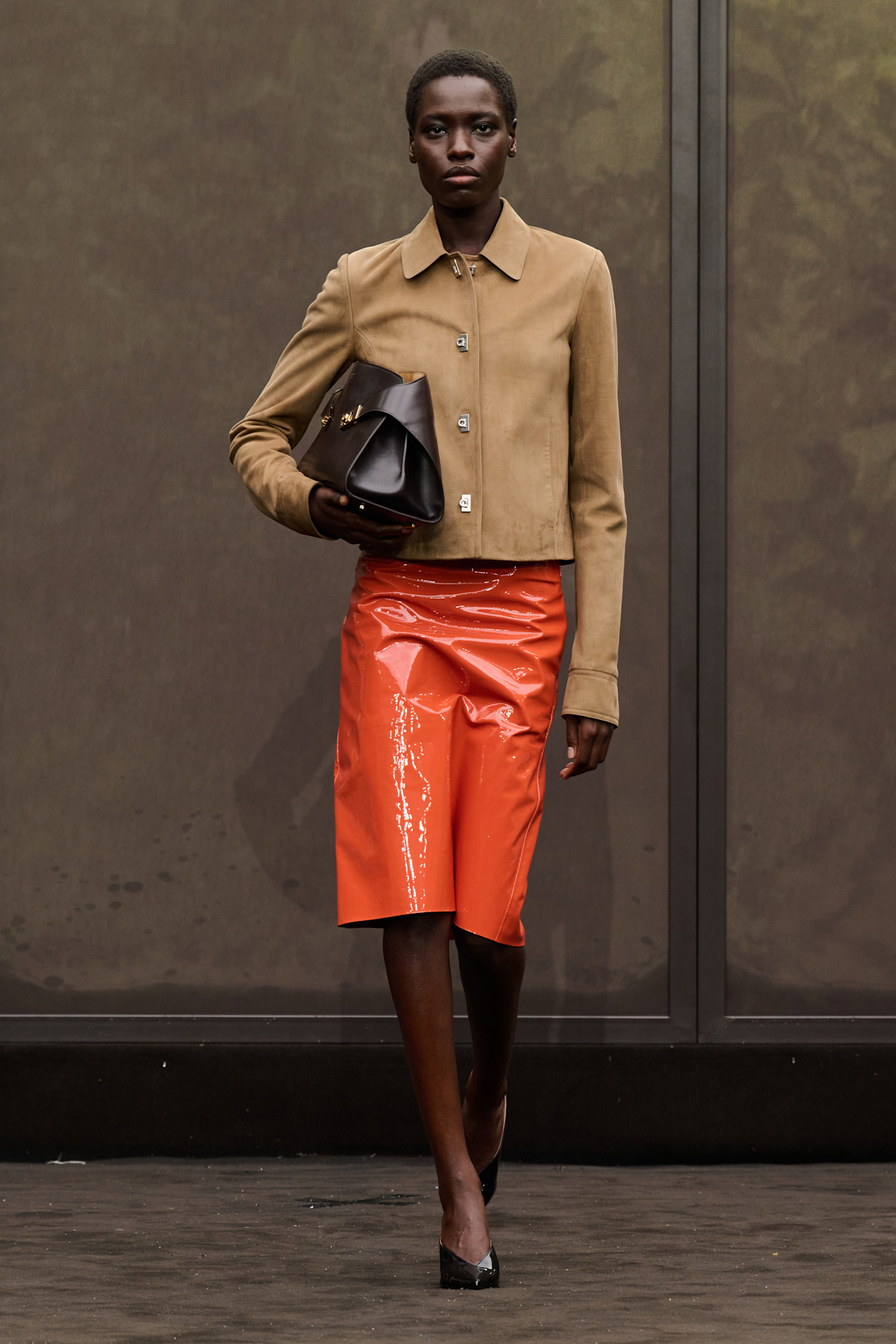 A Ferragamo model wearing a closed light suede jacket with a dark orange patent leather midi skirt, chocolate brown pointed toe mules, and a matching dark brown east-to-west hug clutch bag.