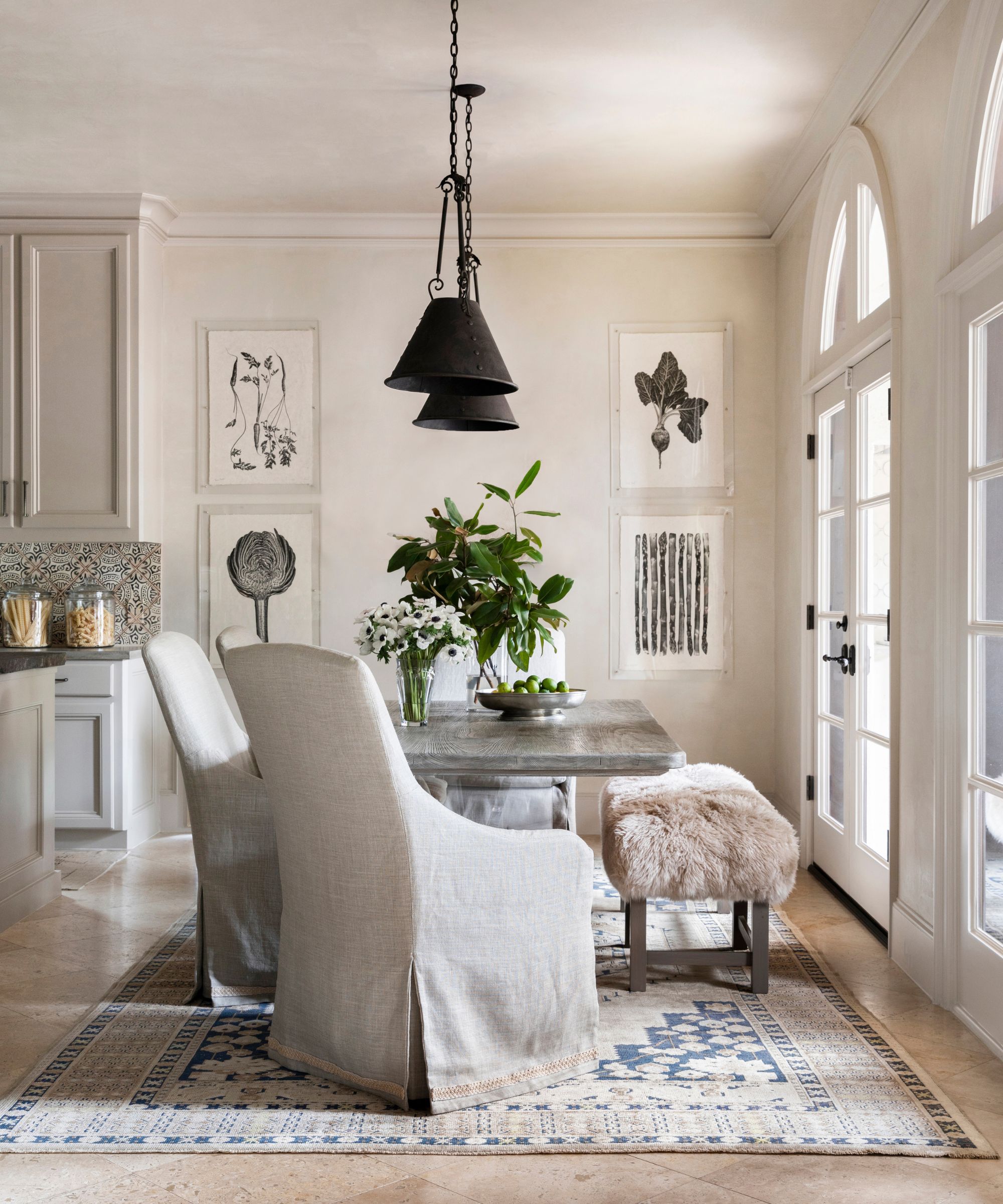 A modern rustic open-plan kitchen with plaster walls, arched windows with French doors, and a large dining area with a large table and upholstered seats, a patterned rug, and two black pendant lights.