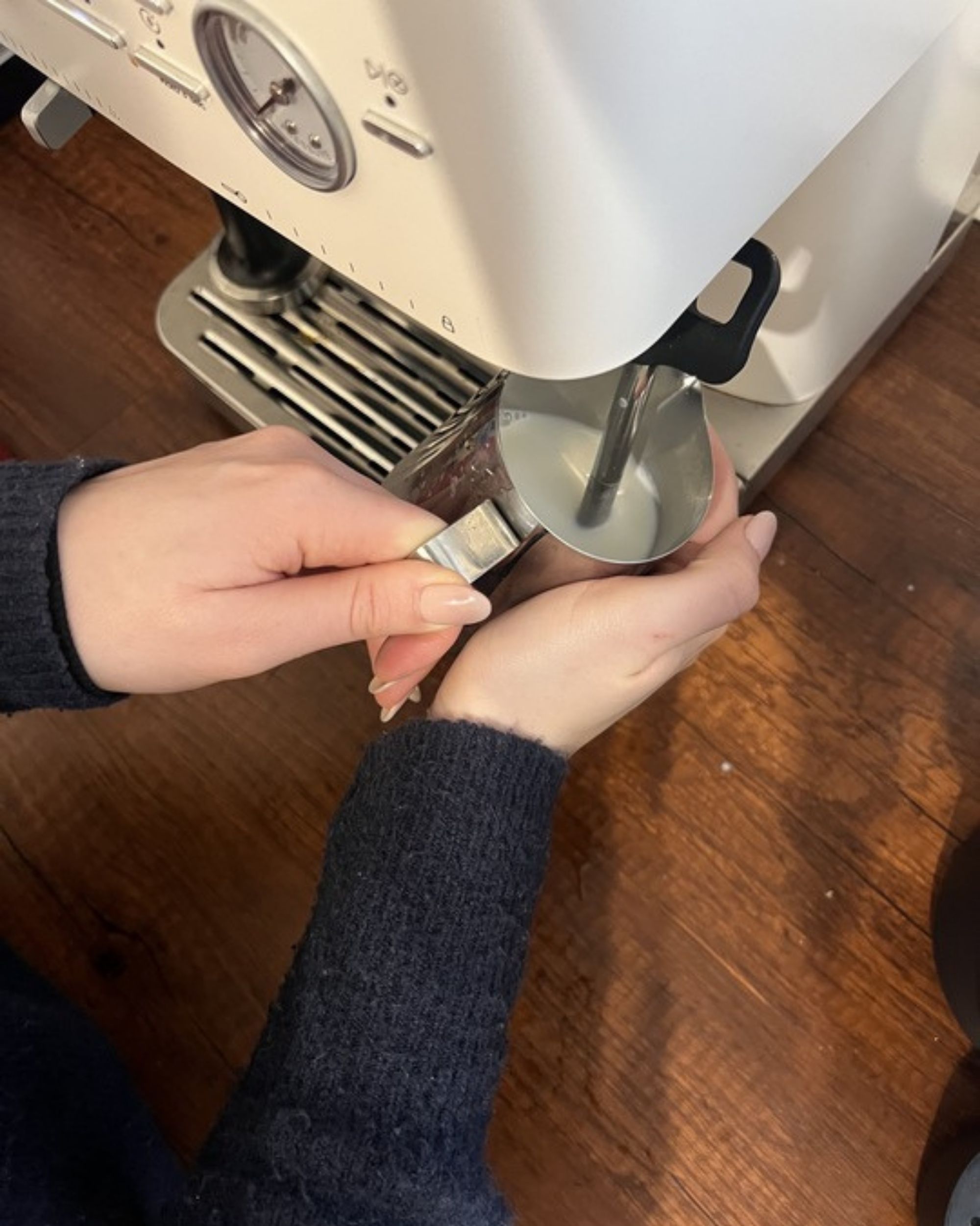 Image of a hand holding a milk jug under a steaming wand attached to a white espresso machine.
