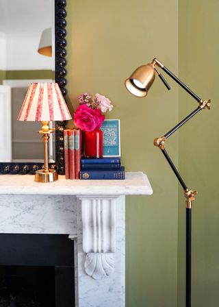 A marble fireplace next to an angle-poise style floor lamp, with a lamp on the mantelpiece with a patterned shade and a brass base