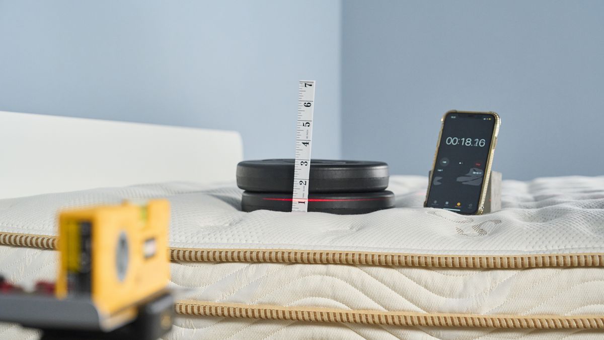 Image shows one of our pressure relief tests in action on the Saatva Classic mattress. A stack of black weights is grouped on top of the Saatva in the shoulder region, with a measuring tape showing how much of an indentation the weight is creating in that area. A stopwatch on an iPhone sits next to the weight stack, so we can time indentation over 5 mins. A yellow laser measure tracks the original starting position of the weights so we have a visual view of indentation too, in addition to the physical measuring tape.
