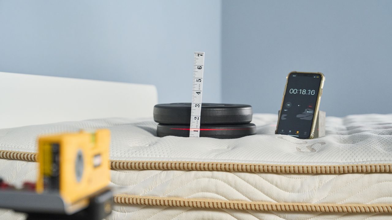 Image shows one of our pressure relief tests in action on the Saatva Classic mattress. A stack of black weights is grouped on top of the Saatva in the shoulder region, with a measuring tape showing how much of an indentation the weight is creating in that area. A stopwatch on an iPhone sits next to the weight stack, so we can time indentation over 5 mins. A yellow laser measure tracks the original starting position of the weights so we have a visual view of indentation too, in addition to the physical measuring tape.