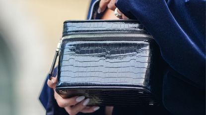 A woman in a chic navy velvet suit is seen holding a black croc vanity case that could be one of the best make-up bags