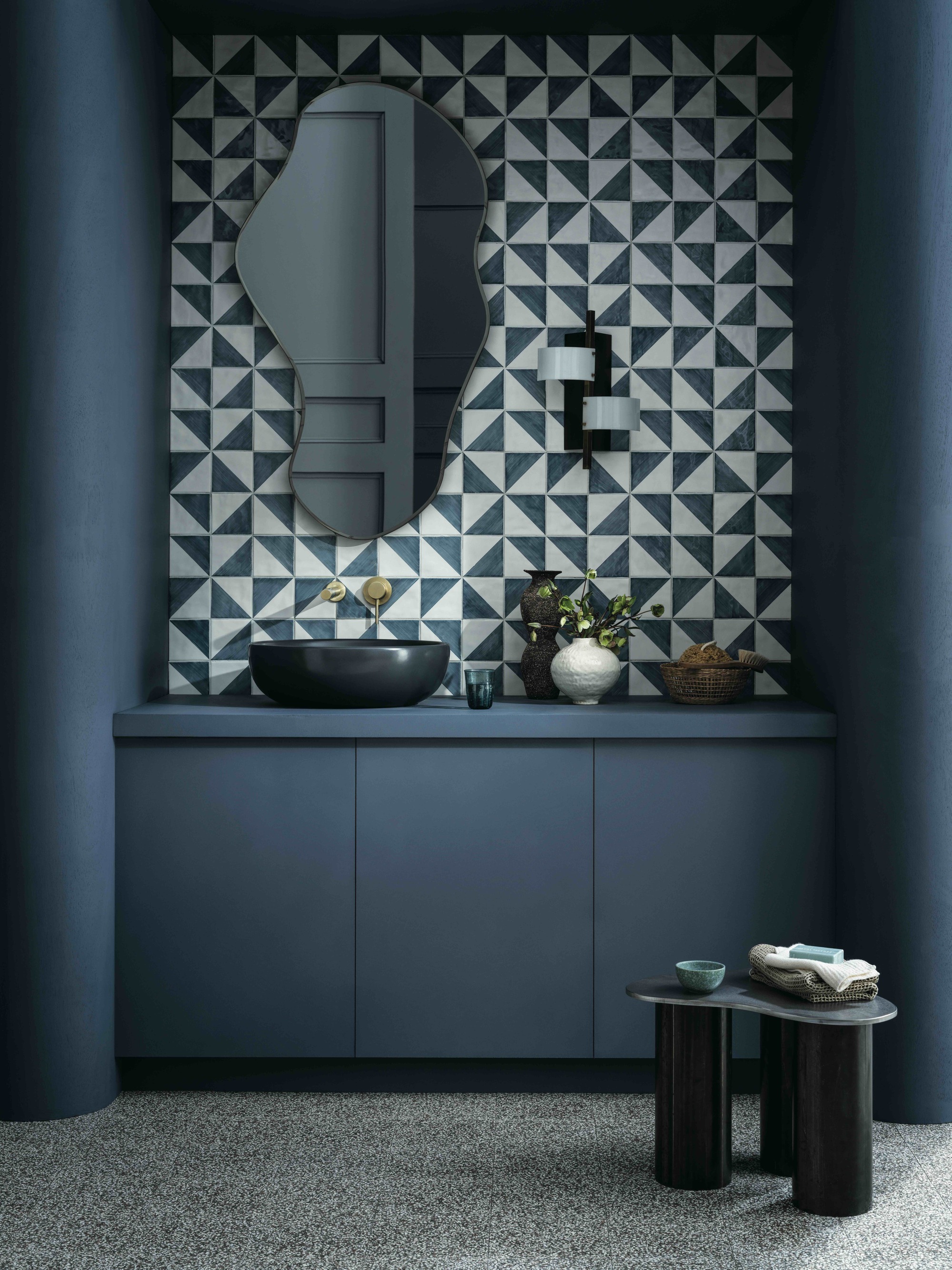 modern blue bathroom with geometric tiles, organic shaped mirror, black stool with cups and bath towels, terrazzo floors