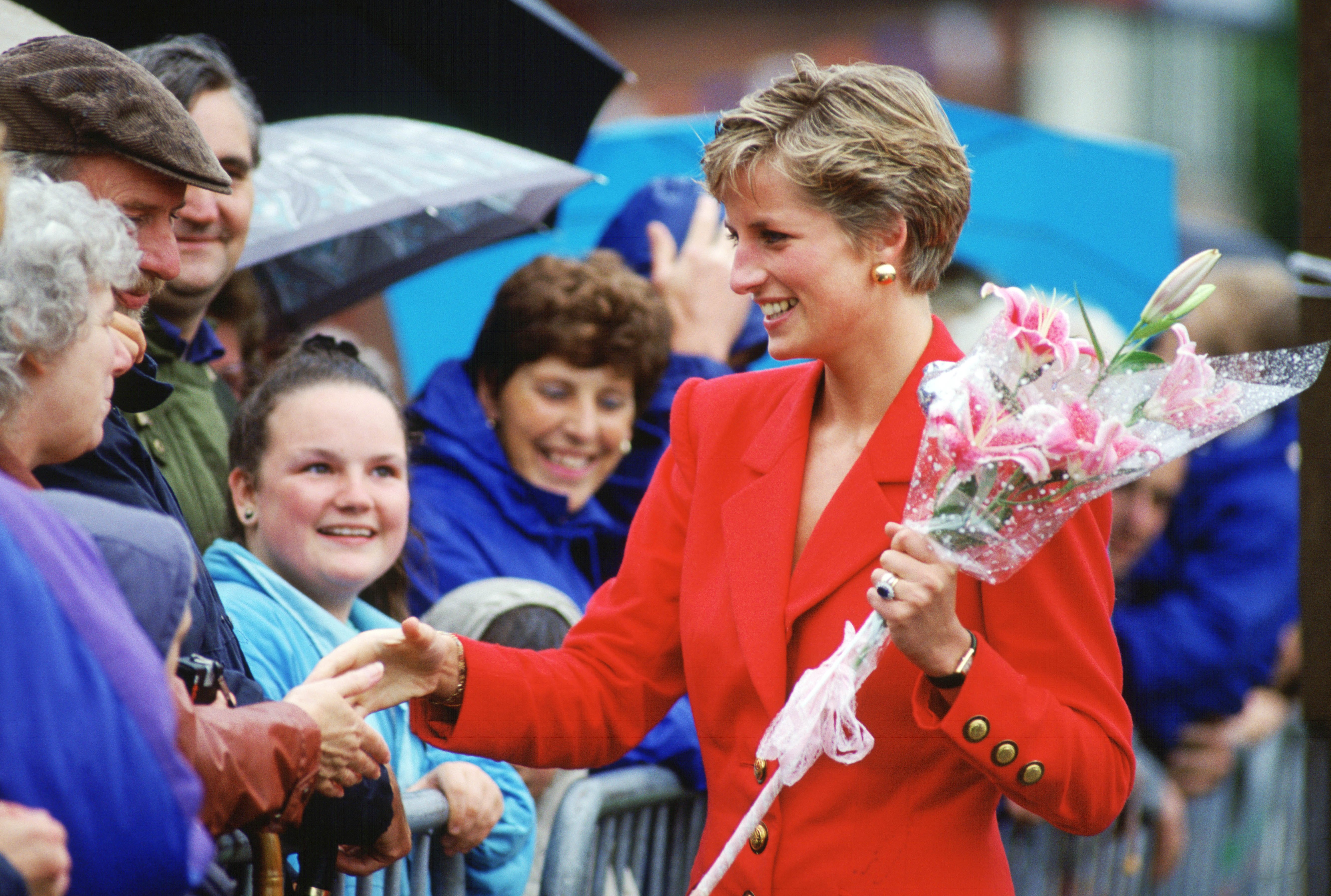 Princess Diana wearing a red jacket shaking hands with fans