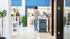 Man using a printer in an open plan office space while talking to colleague.