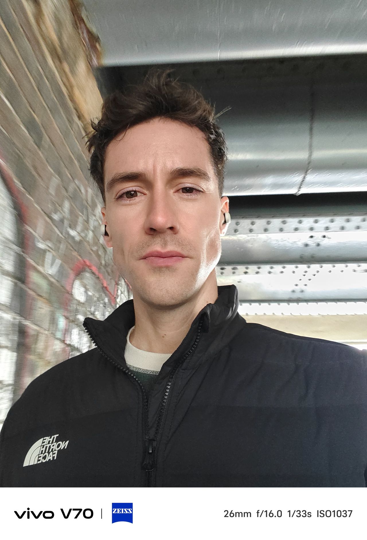 Indoor selfie of man under bridge with metal beams and brick wall behind.