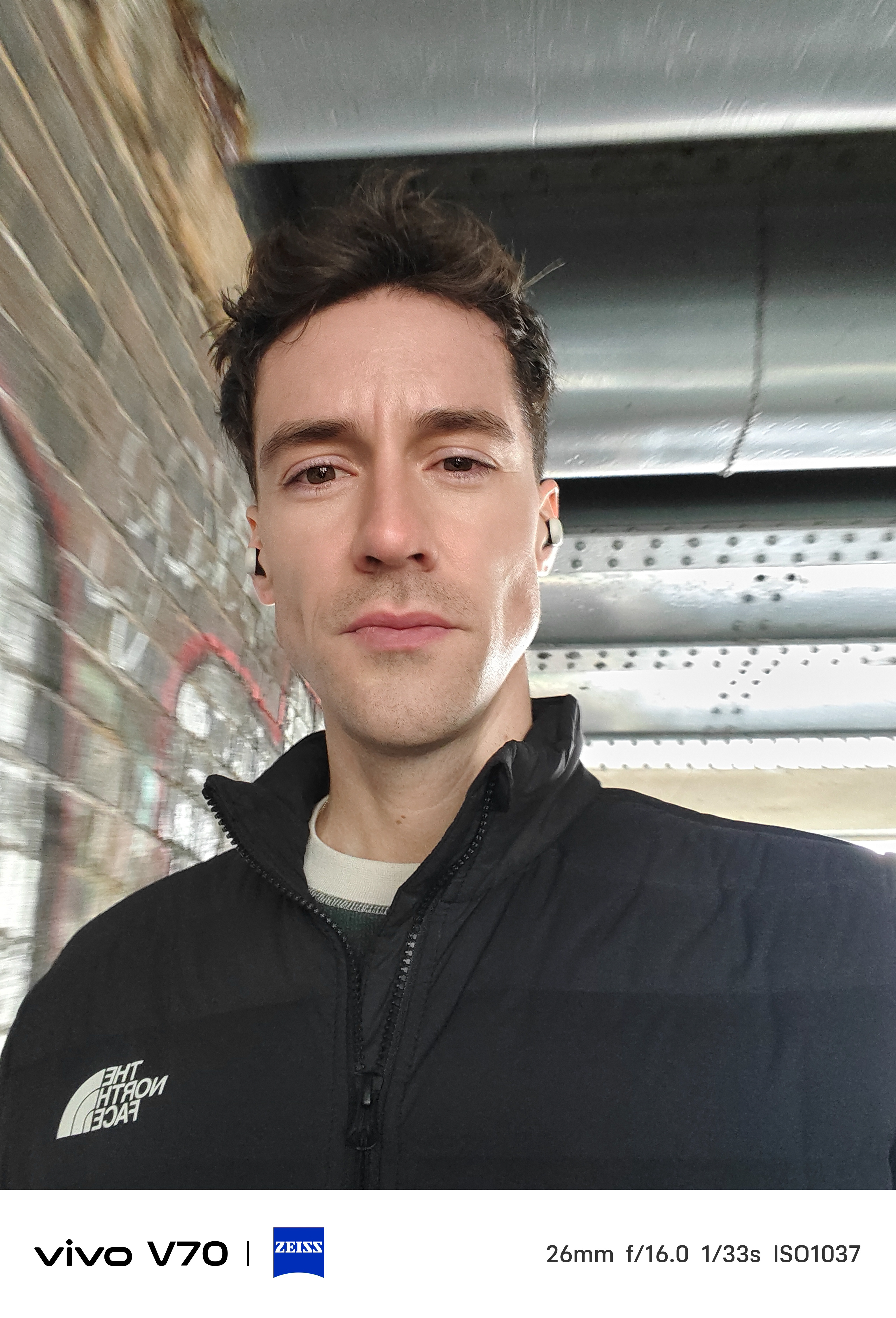 Indoor selfie of man under bridge with metal beams and brick wall behind.