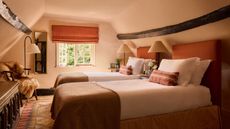 A loft bedroom with sloped ceilings color-drenched plaster pink, an exposed wood ceiling beam, and twin beds with terracotta headboards, crisp white bedding and terracotta cushions and throws. 