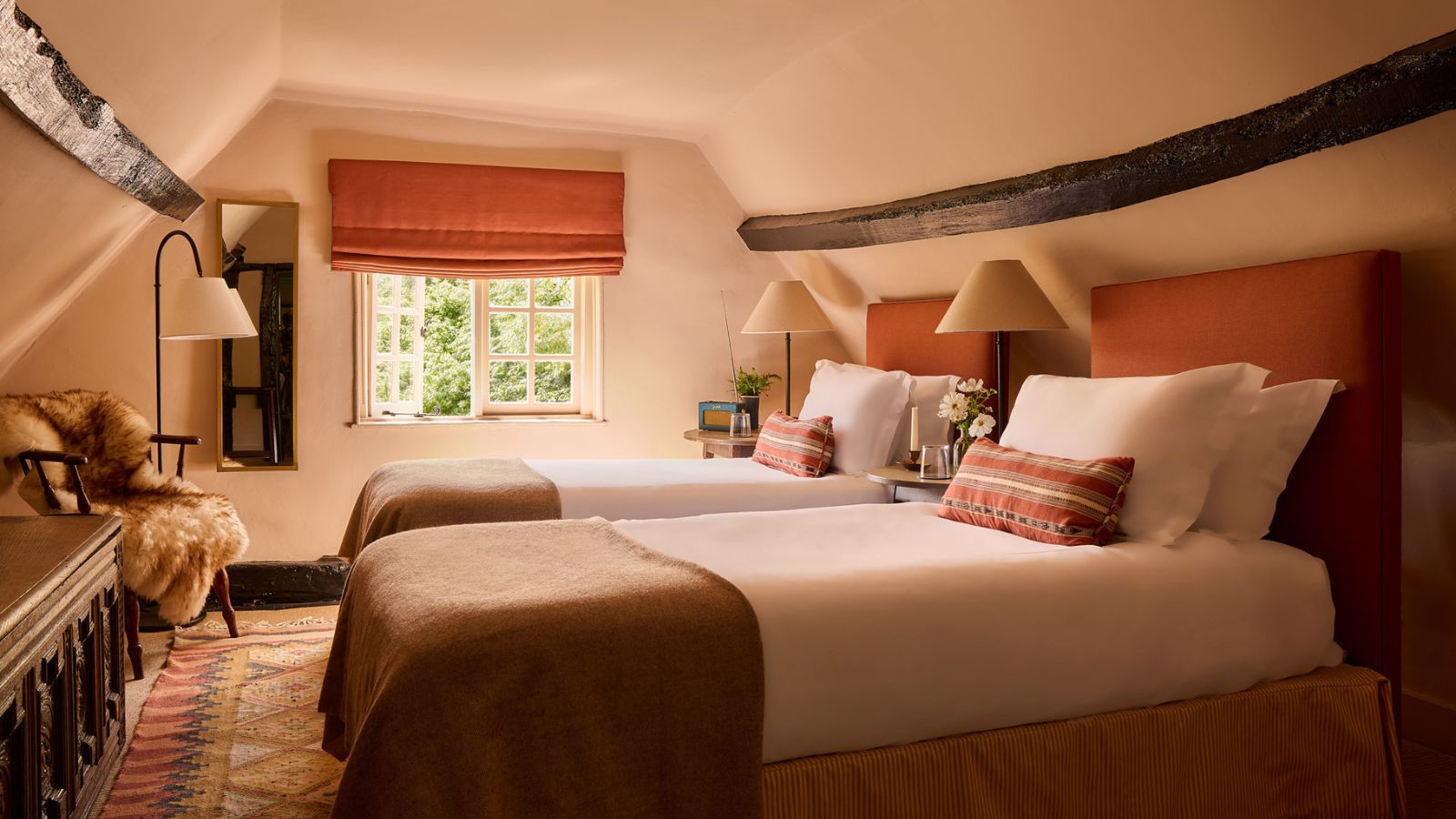 A loft bedroom with sloped ceilings color-drenched plaster pink, an exposed wood ceiling beam, and twin beds with terracotta headboards, crisp white bedding and terracotta cushions and throws. 