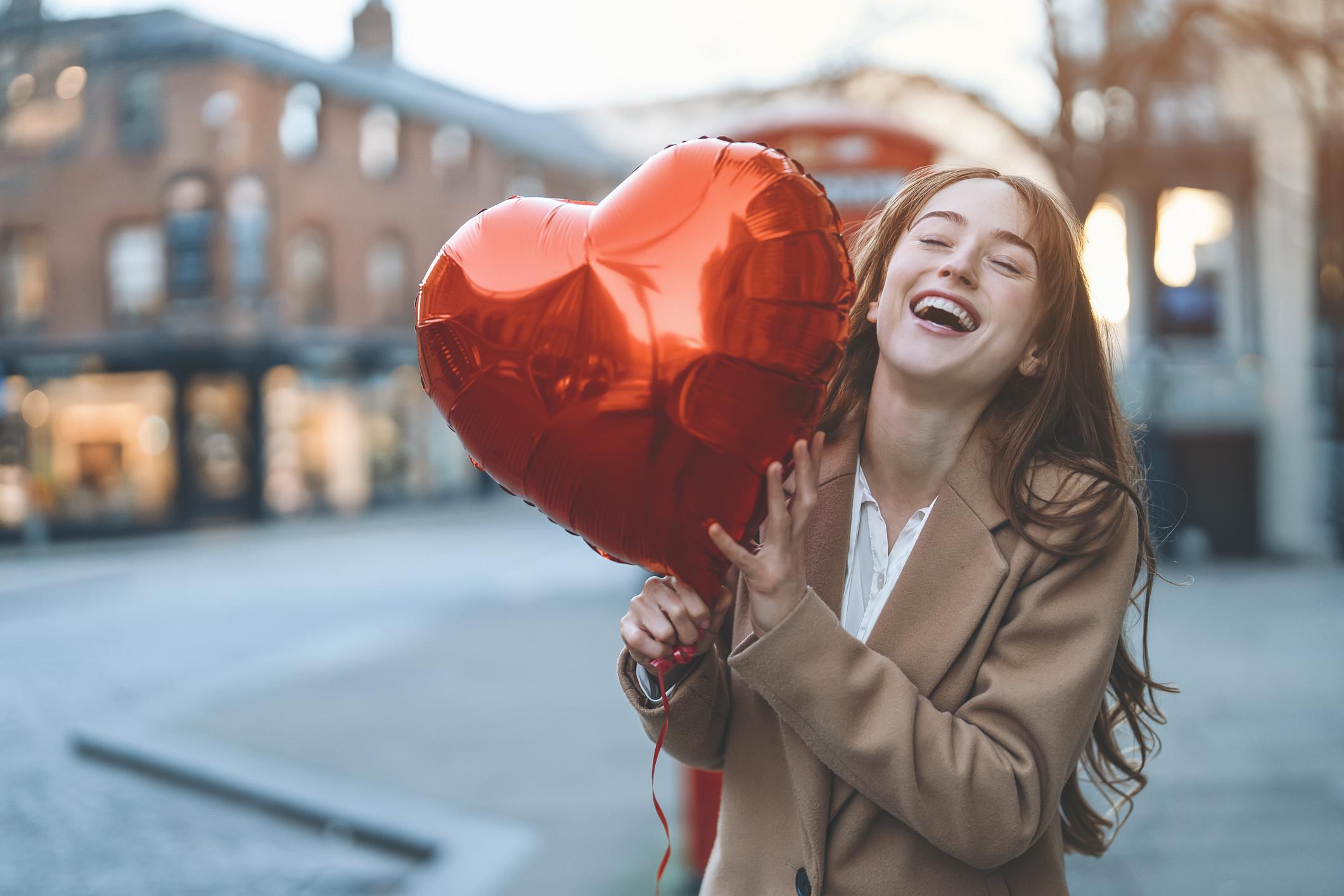  A woman with long brown hair is smiling, she is holding a large red heart balloon in the street.  