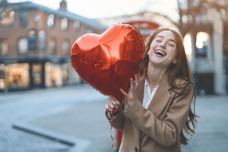 A woman with long brown hair is smiling, she is holding a large red heart balloon in the street. 