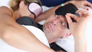 A man lifting his sleep mask off his face and making a surprised face at his wife's sleep talking who's lying next to him