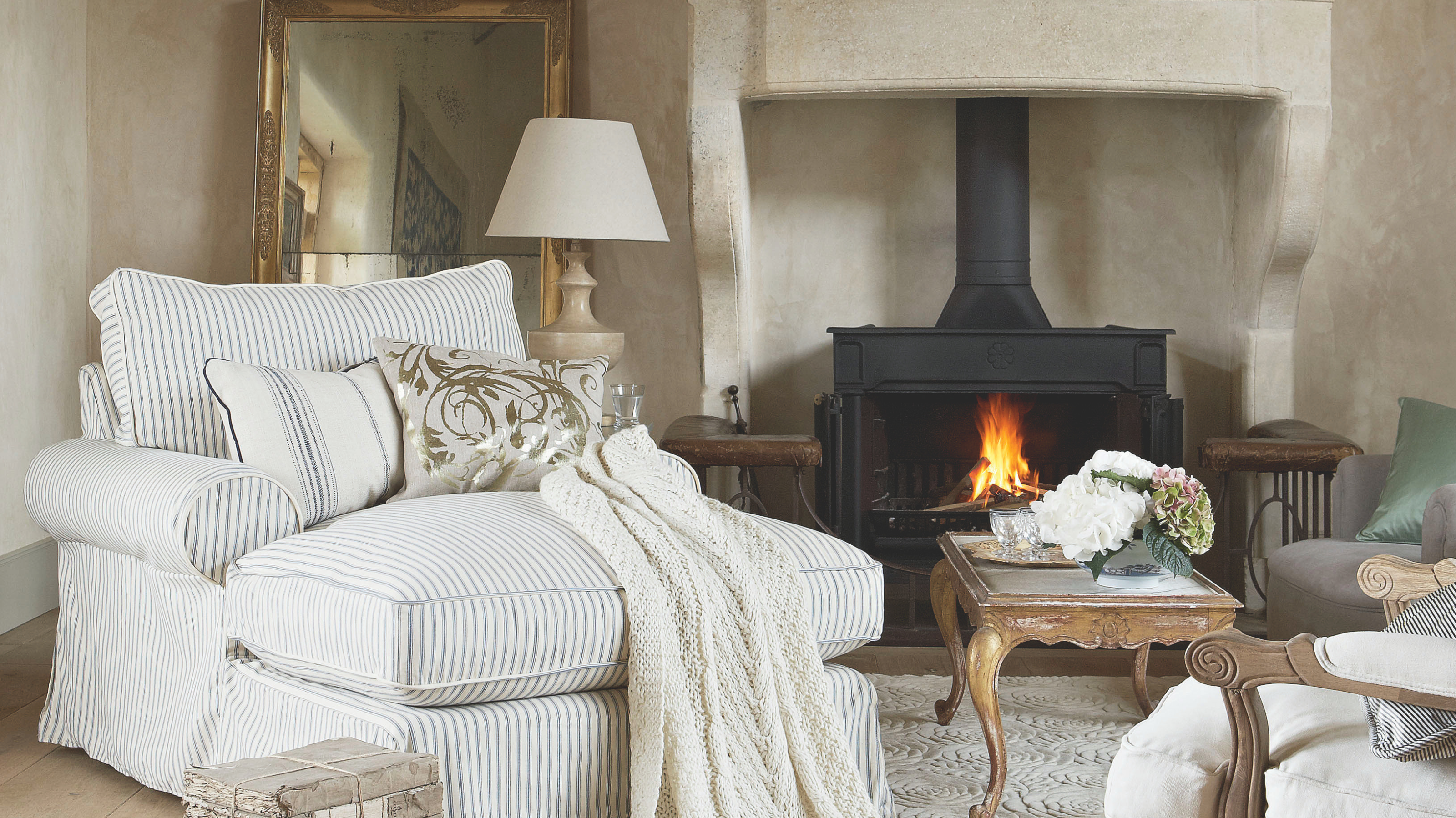 Neutral cosy living room, stone fireplace, lit stove, giant oversize clock face, grey striped armchair, gold gilded coffee table, wood flooring