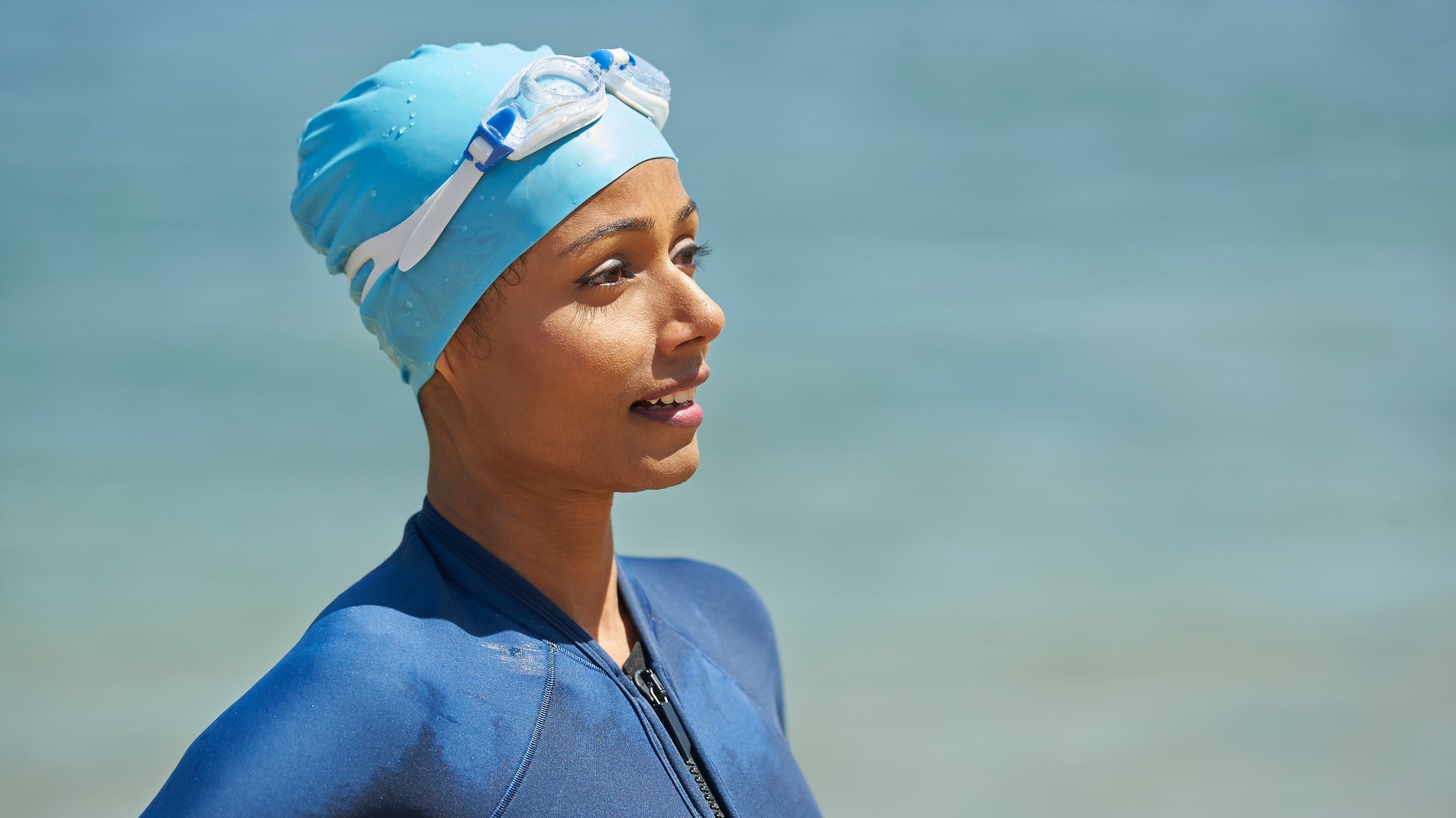 A swimmer stands in front of a natural body of water. She wears a swim cap on her head, and she wears a swimsuit. Both are different shades of blue.
