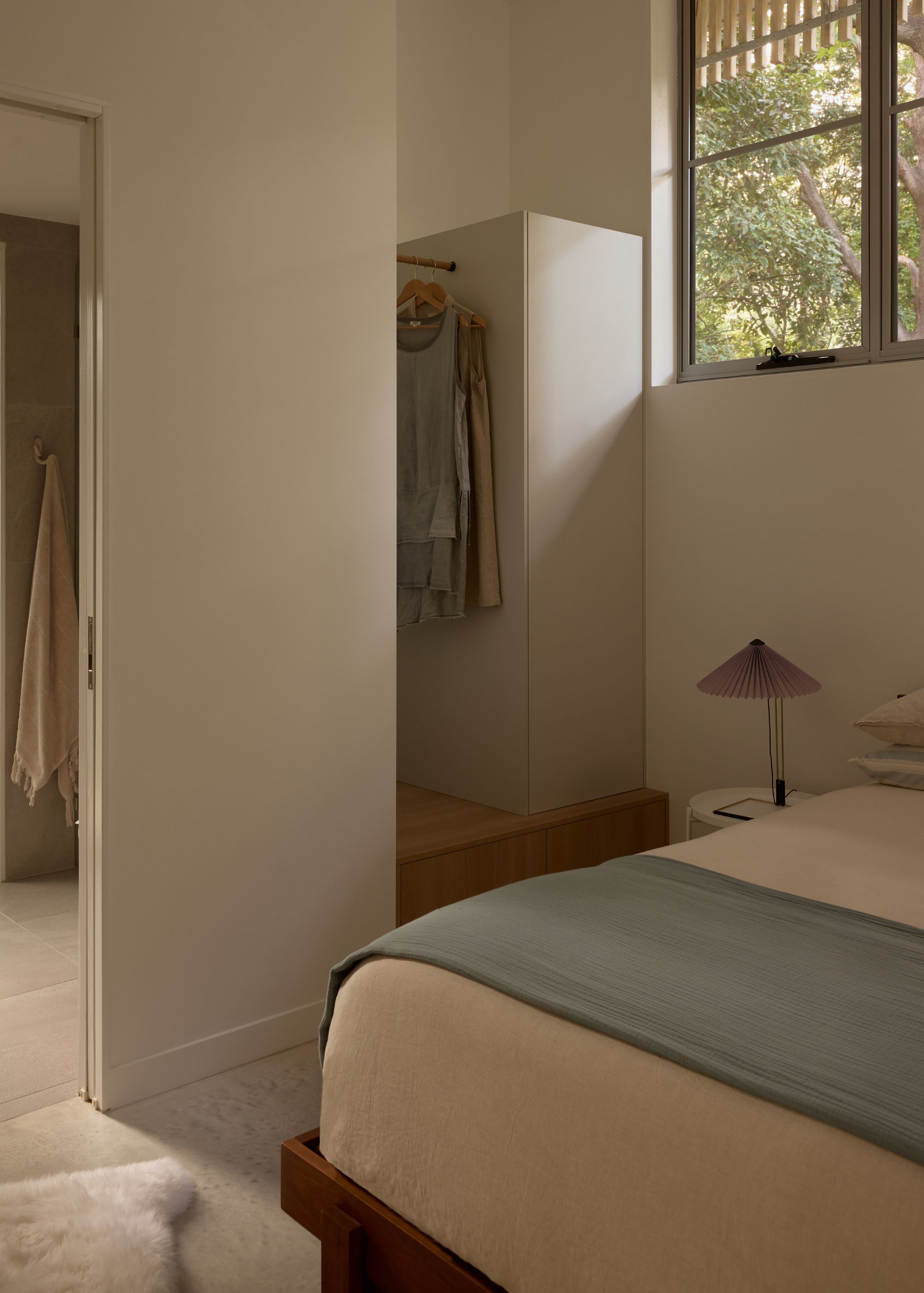 A white bedroom with a wardrobe, a pleated table lamp, and a blue coverlet on a cream bedscape