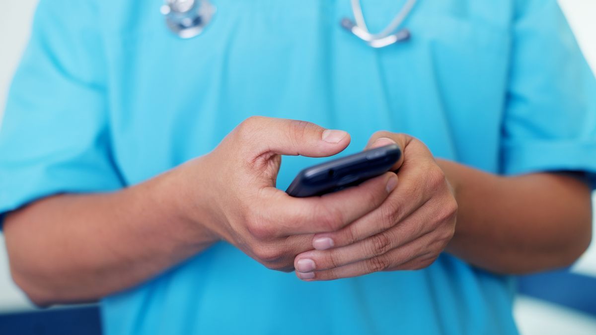 A doctor in blue scrubs holds a phone in both hands, with only their arms, midriff and chest visible