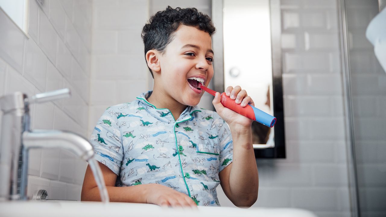 A young boy standing in front of a sink using one of the best children&#039;s electric toothbrush