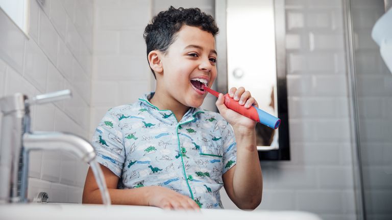 A young boy standing in front of a sink using one of the best children's electric toothbrush