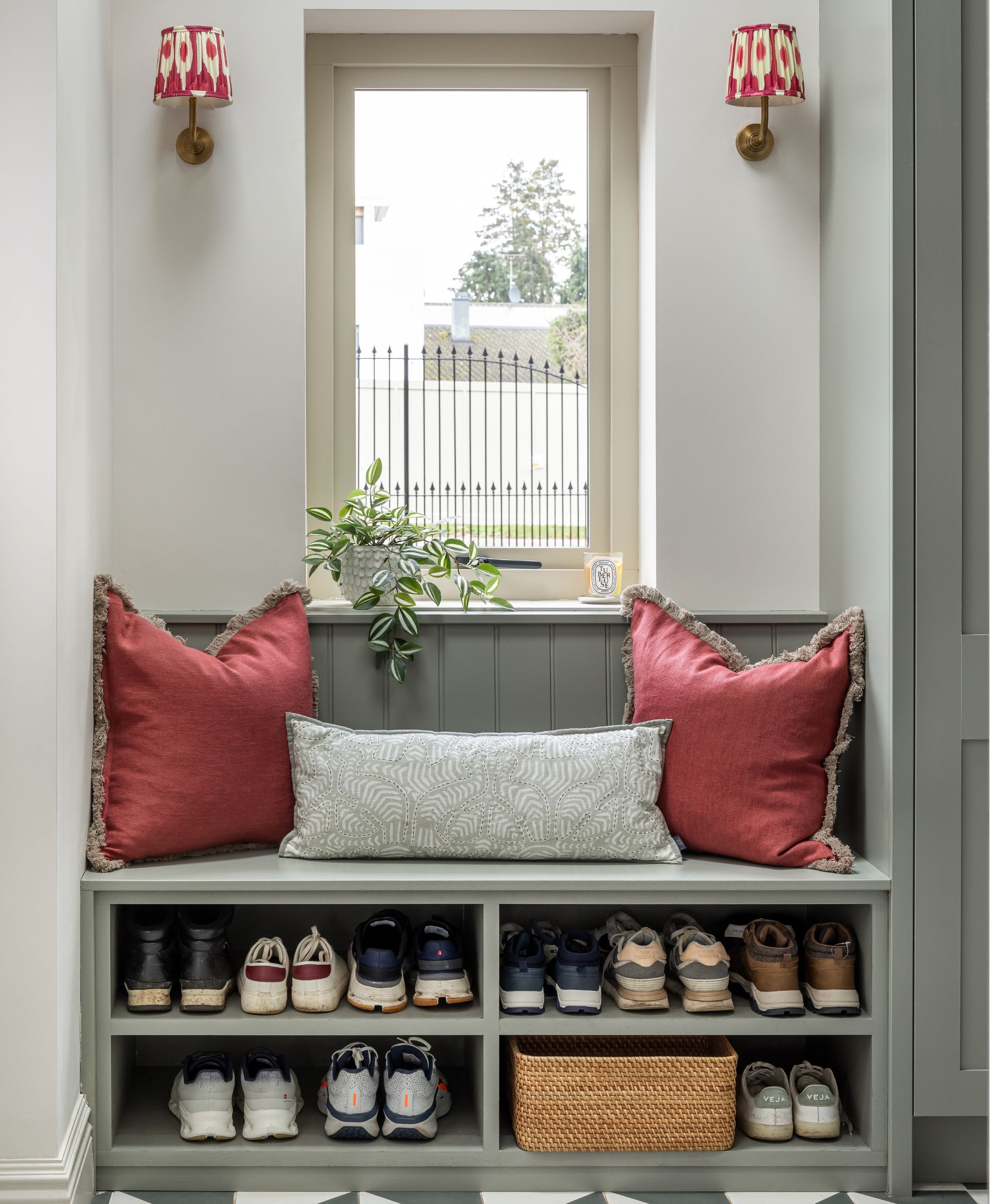 A sage green built-in hallway bench with open shoe cubbies below holding multiple pairs of trainers and a wicker basket. Two pink cushions with fringe trim and a patterned bolster cushion sit on the seat, flanked by brass wall sconces with red ikat shades.