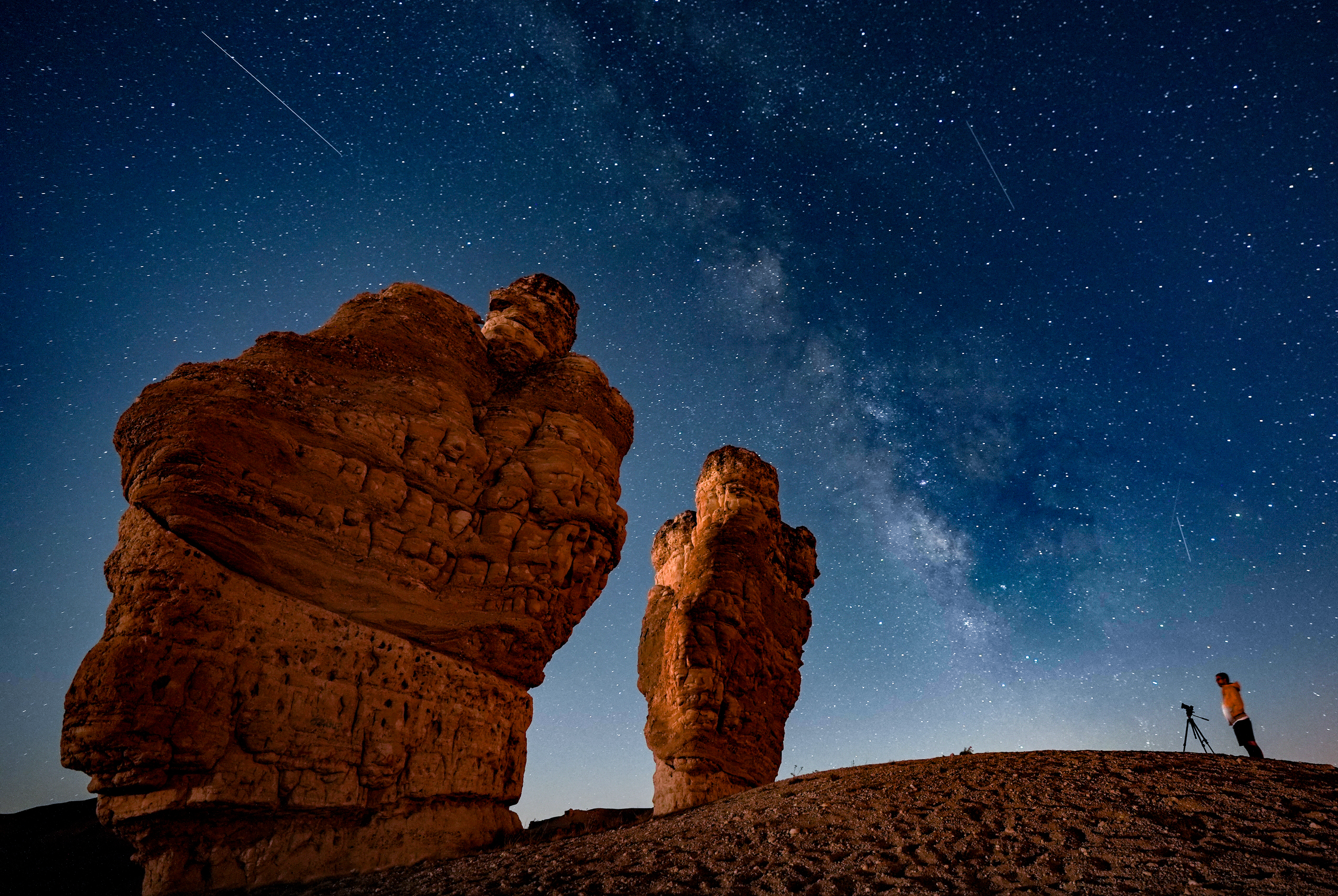 shooting stars can be seen running alongside the glowing band of the Milky Way above two tall rock formations. A man with a camera is visible to the left standing on a sandy hillock.