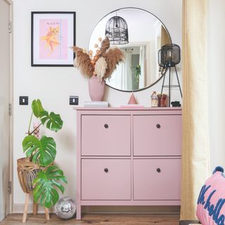 A hallway with a pink shoe cabinet and a round mirror on the wall above it