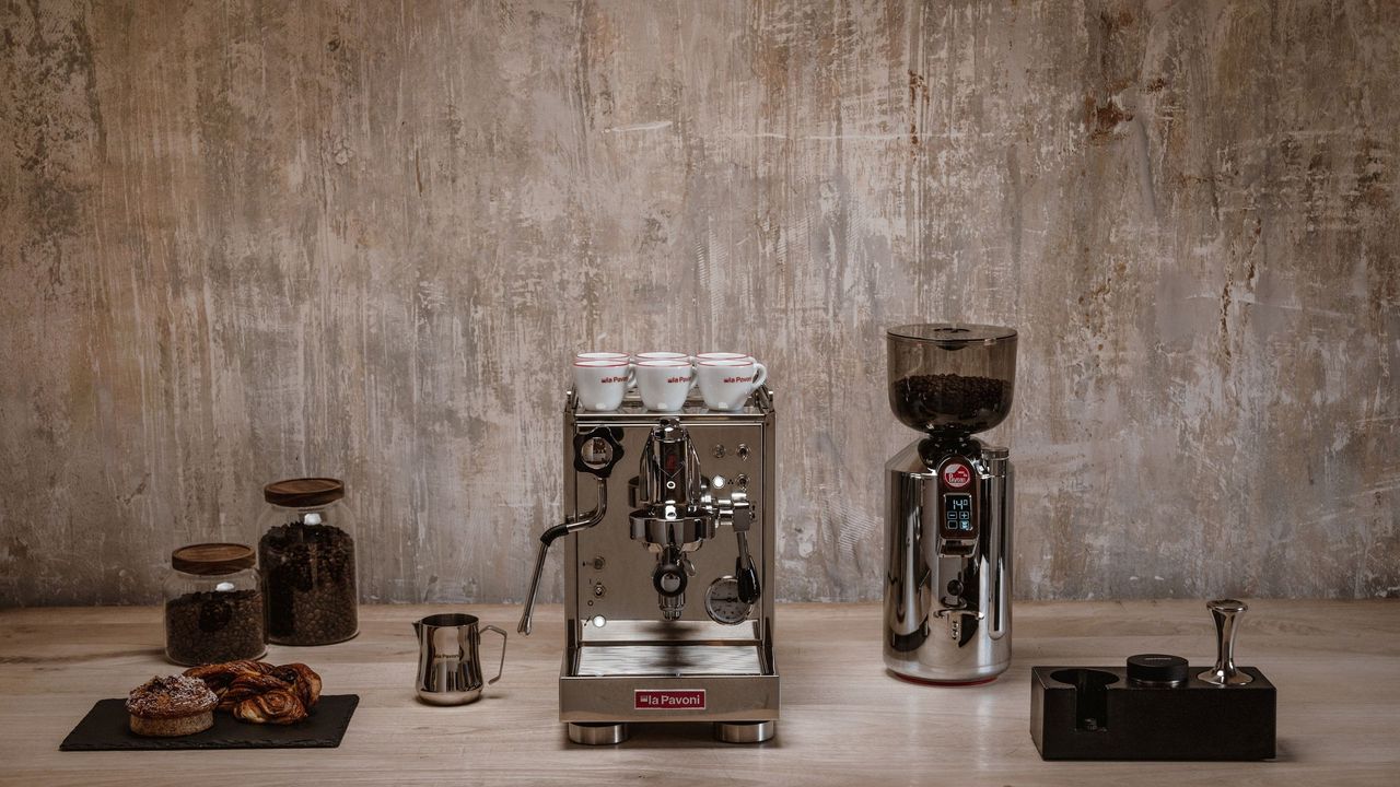 Image of a stainless steel La Pavoni machine on a beige countertop with a beige wall behind it. There are other coffee machine accessories around it.