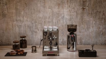 Image of a stainless steel La Pavoni machine on a beige countertop with a beige wall behind it. There are other coffee machine accessories around it.